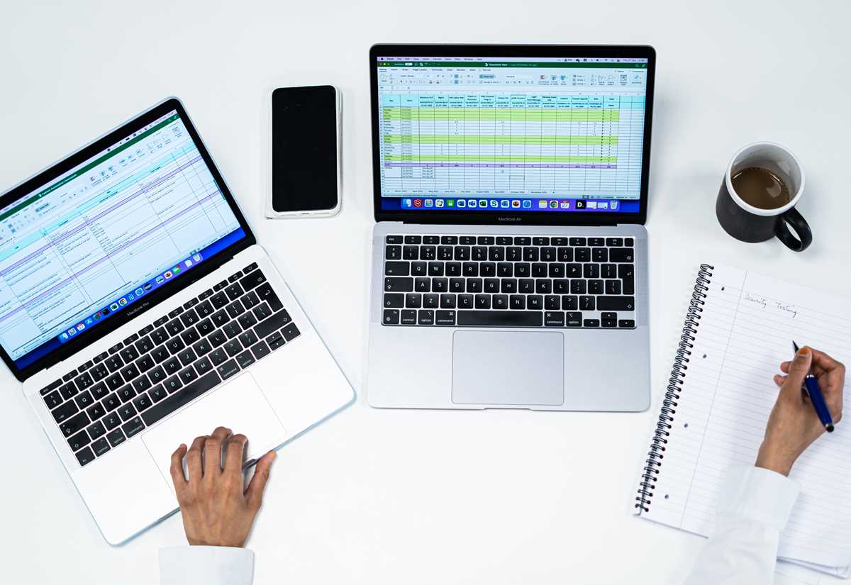 Overhead view of two laptops showing spreadsheets, a phone and a coffee, with a person writing in a notebook