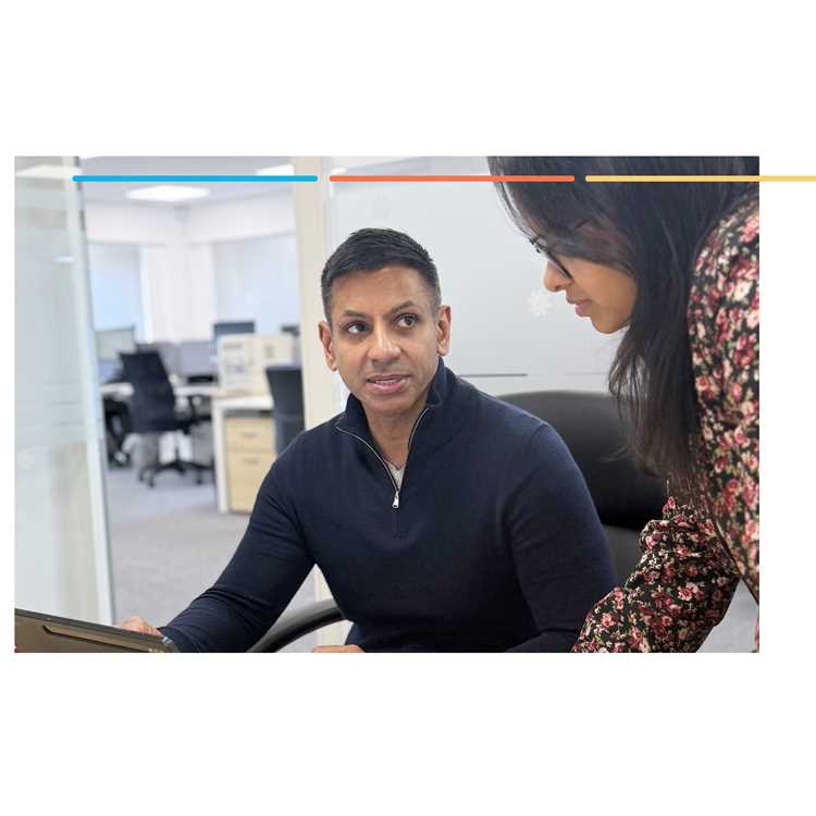 A man in a navy zip-neck jumper looks at a laptop in an open-plan office while a woman stands beside him, also looking at the screen
