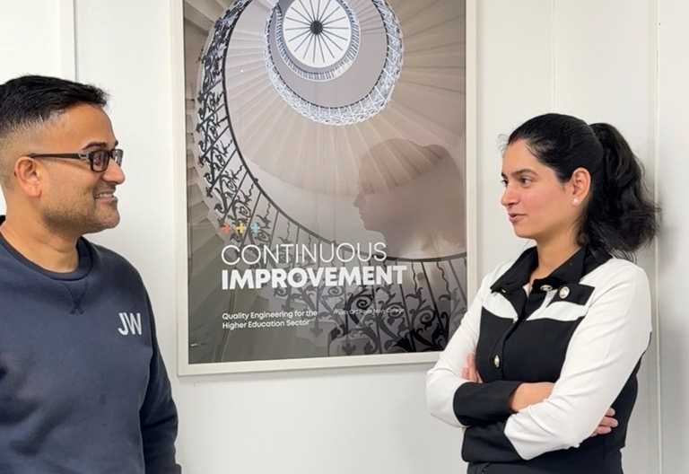 A man and a woman standing either side of a framed Prolifics Testing Continuous Improvement poster, talking