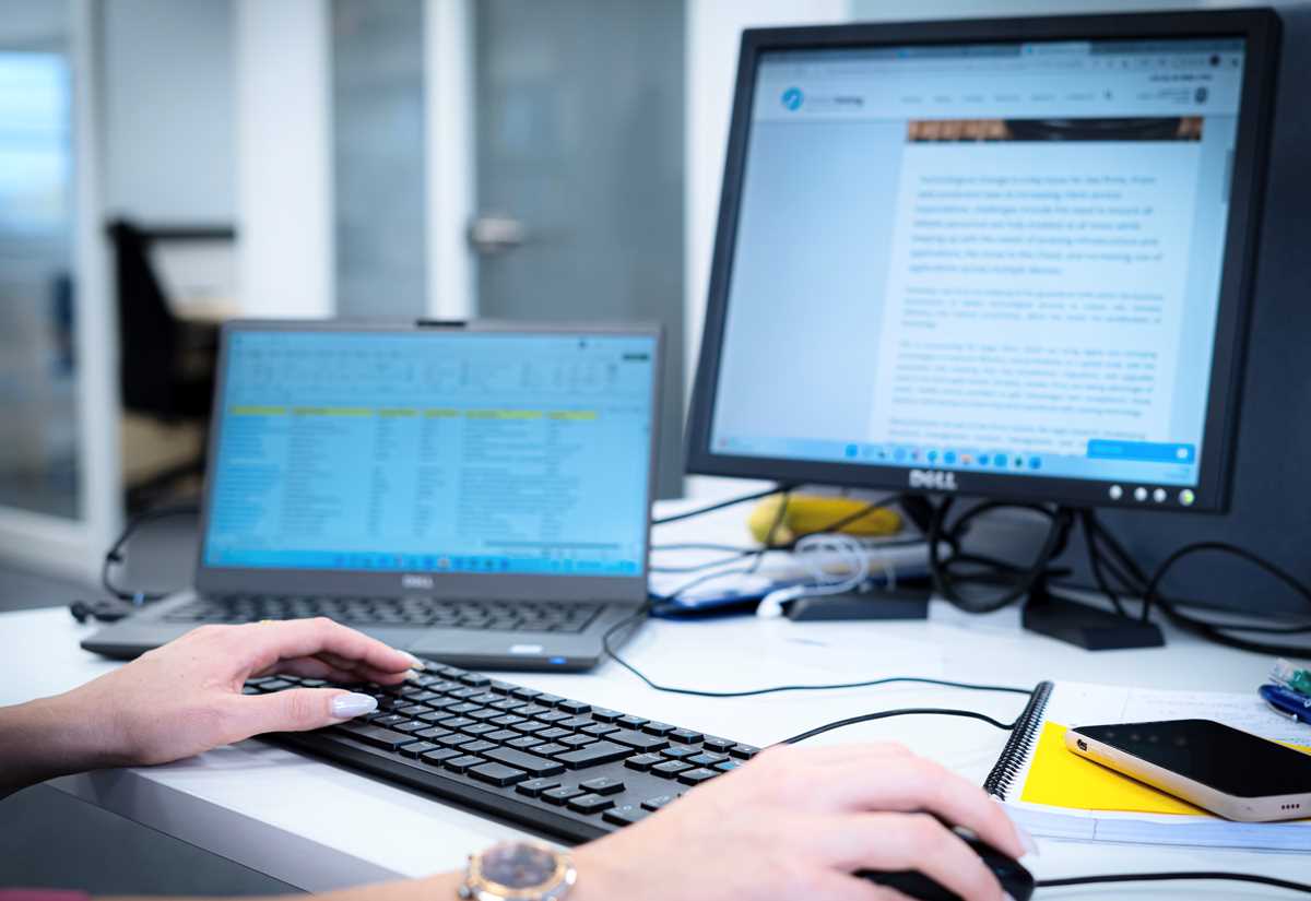 Hands typing on a keyboard and using a mouse at a desk with a laptop, two monitors and a phone